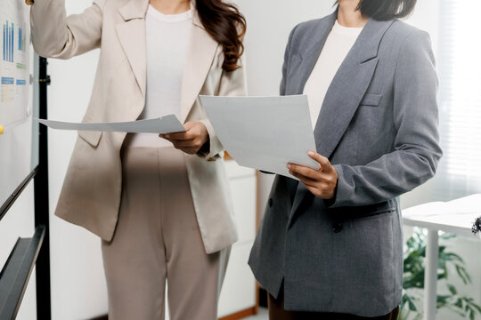Two businesswomen discussing financial data and charts on a whiteboard, working together in a professional office environment - Powered by Adobe