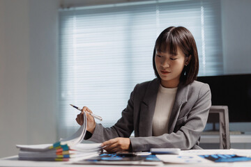 Young Asian businesswoman working in office, reviewing documents and financial data on her desk, focused on professional tasks