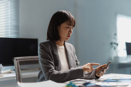 Asian businesswoman working at desk, analyzing financial data and using smartphone for communication and business tasks