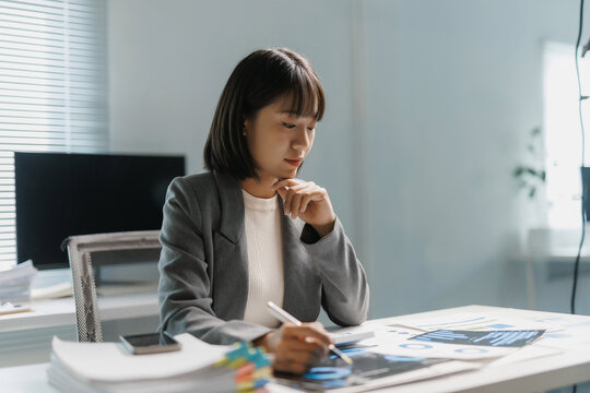 Young professional Asian businesswoman working at desk, analyzing financial data, charts, and reports for business strategy - Powered by Adobe