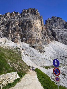 Start of the Tre Cime hiking trail circuit at Rifugio Auronzo, with Il Mulo on the left and Cima Grande on the right; hikers on the path below sheer limestone cliffs under blue sky