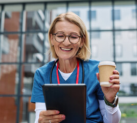Nurse in scrubs enjoying coffee and using a tablet computer  outside a modern medical facility during a sunny day
