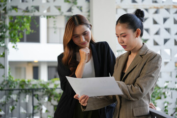 Two Asian businesswomen collaborating, reviewing, and analyzing paper documents together outdoors in a professional setting
