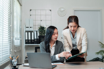 Two professional businesswomen collaborating, reviewing documents, and discussing work while using a laptop in a contemporary office setting