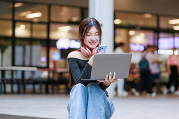 Young woman smiling, holding a laptop and smartphone, engaging with technology, wearing headphones in a casual setting