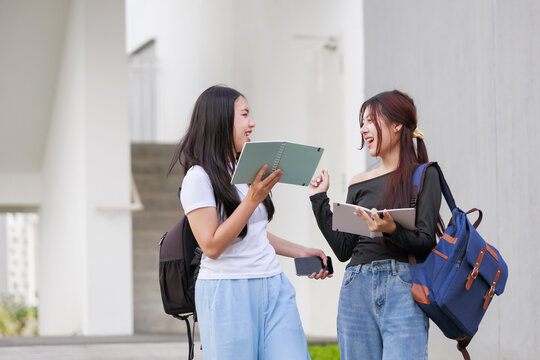 Two Asian female students discussing notes, laughing together outside university, carrying backpacks and digital devices