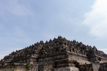 Fototapeta premium An aerial view of the 9th-century Borobudur Temple, a UNESCO World Heritage site built of andesite stone. The Buddhist monument is surrounded by lush tropical trees under a bright morning sky.