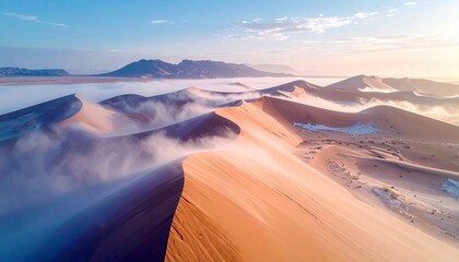 Aerial view of sand dunes with mist and mountains in the background during sunrise. The scene captures the beauty of nature and the desert landscape.