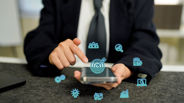 Business Goals, A businessman holding a smartphone with a glowing target icon, surrounded by other business icons for charts, ideas, and gears - Powered by Adobe