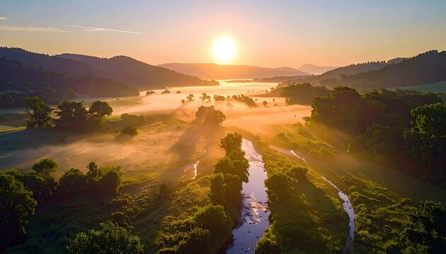 Aerial view of a river valley at sunrise, with fog, trees, and sun rays creating a warm, golden atmosphere.