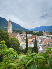 Cityscape of Merano in South Tyrol, Italy with St. Nicholas Church and bell tower above rooftops; green hills and distant mountains under a sky with clouds