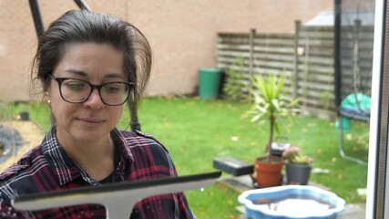 Woman cleaning windows in garden wearing glasses and plaid shirt during the day