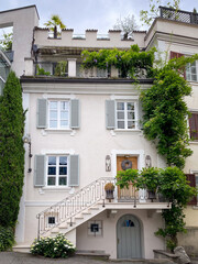 Charming townhouse facade with pastel walls, shutters and arched door; staircase with wrought-iron railing and lush balcony/roof-terrace plants under a sky with cloud in Italy