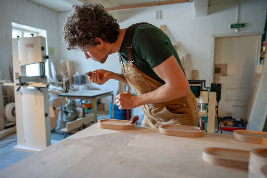 Man carpenter working on set of coasters in carpentry workshop, blows dust and sawdust off chisel blade, sanding coasters for water glasses with sand paper, working process of small woodshop business.