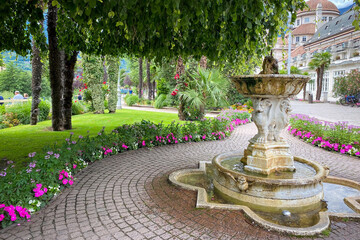 Ornate stone fountain with flowerbeds and palms on the Passer Promenade in Merano (Meran), Italy; landscaped paths near the Kurhaus under a sky with cloud
