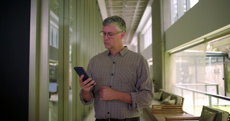 Middle aged man standing in modern office hallway using cellphone with focused expression browsing screen and interacting with mobile device in business setting