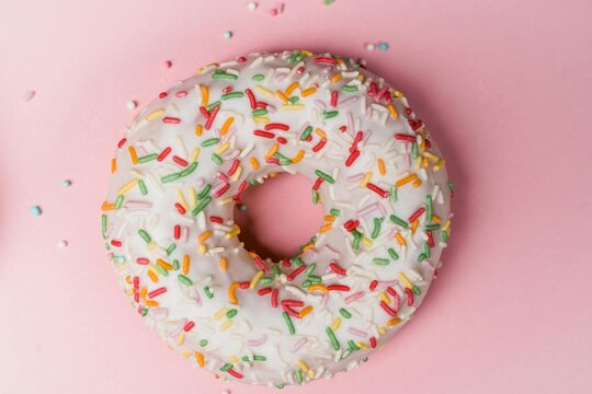 Overhead view of a donut with white frosting and colorful sprinkles on a pink background surface - Powered by Adobe