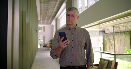 Middle aged man standing in modern office hallway using cellphone with focused expression browsing screen and interacting with mobile device in business setting