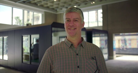 Middle aged man standing in office hallway smiling at camera in confident relaxed posture bright modern professional business environment