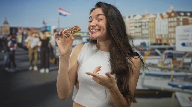 Woman holds granola bar in hand with wide open mouth gesture at building where boats dock under clear sky; joy.