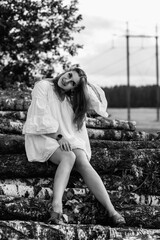 Young woman in a flowing white dress sits gracefully on stacked logs, surrounded by nature, with a serene expression and soft lighting, embodying tranquility and beauty