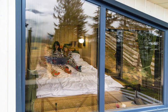 Family in cabin watching laptop through window. Happy family in matching pajamas relaxing on bed inside cozy cabin, watching laptop together. Seen through window with outdoor reflections.