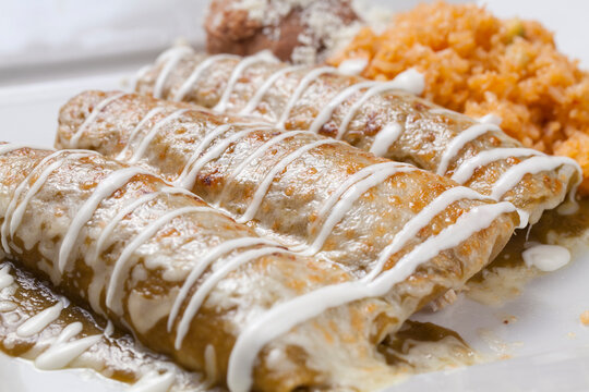 Grilled Mexican chicken enchiladas with white cream, green sauce, beans and rice on a white plate. 