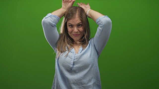 Young blonde woman making ear gesture with hands on head in green studio setting, casual blouse and shell necklace; playful confidence.