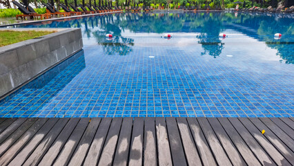 Close-up view of a swimming pool with blue mosaic tiles and wooden deck, surrounded by sun loungers and greenery, creating a relaxing tropical resort atmosphere.