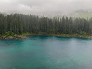 Rain softens the turquoise water of Lake Carezza (germ. Karersee) beneath spruce and fir forest in South Tyrol, Italy, with low mist drifting across the Dolomites