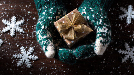Hands in festive green mittens hold a gold-wrapped gift box amidst scattered snow and snowflakes, perfect for winter holidays or Christmas.