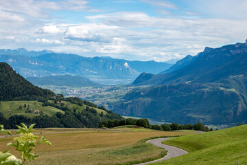 Obraz premium Rolling pasture above the Adige Valley looking toward Ueberetsch (Oltradige), south of Bolzano; winding street and distant mountains under blue sky with cloud