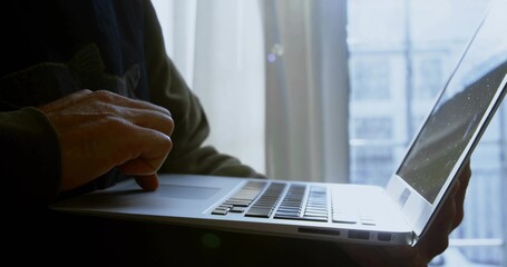 Typing mature woman holding silver laptop at window with sheer curtain, wearing dark long sleeves