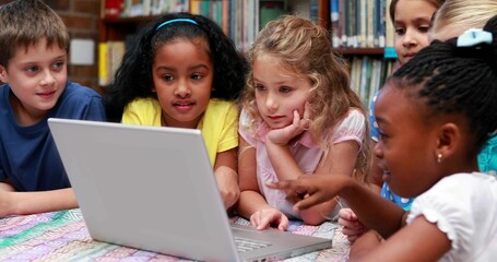 Leaning group of six students pointing at silver laptop on patterned table in library with shelves