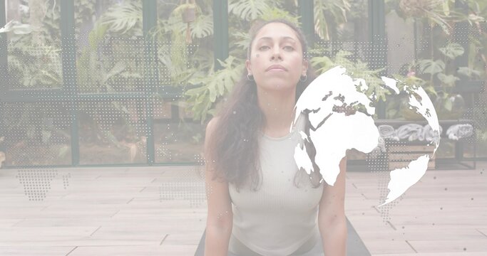 Arching woman doing yoga backbend on mat in glass patio, wearing light-gray tank with globe-overlay