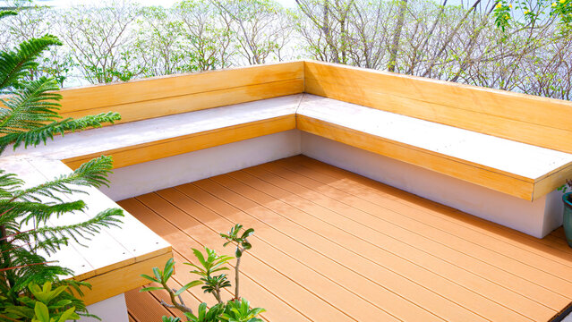 Terrace area of modern house with built-in light wood bench seating and warm brown composite decking under bright daylight, surrounded by lush green foliage, perspective side view with copy space
