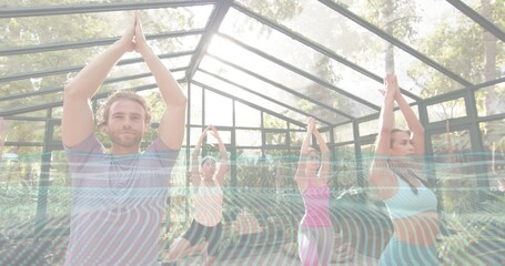 Leading man in striped T-shirt holding balancing pose inside glass greenhouse, with yoga mats