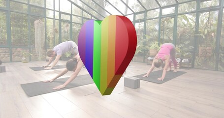 Practicing yoga trio holding down dog on yoga mats in glass room, activewear, rainbow heart, block
