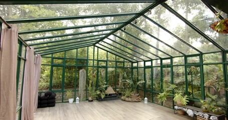 Displaying green metal-framed conservatory, showing pitched glass roof, potted plants, cushions