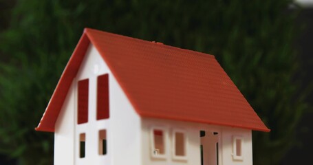 Showing white scale model house sitting on flat table near blurred green plants, featuring red roof