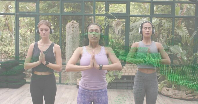 Praying trio of women in athletic wear in glass studio, wearing headphones, watch, stacked mats