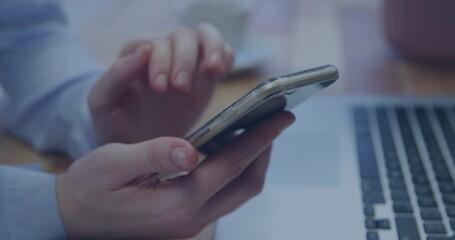 Tapping hands wearing light-blue cuff, holding smartphone at small home desk with laptop and mug