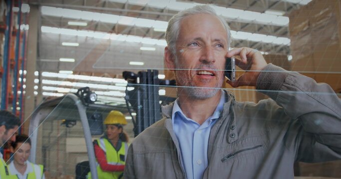 Talking manager on phone in warehouse behind glass partition, with forklift and vests