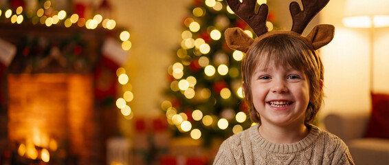 Christmas joy of a child: a smiling child wearing festive reindeer antlers radiates pure joy, set against a backdrop of a warm fireplace and illuminated Christmas tree. Christmas banner