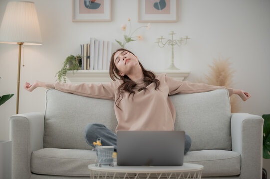 Relaxed serene pretty young Asian woman stretching body for relaxing on comfortable sofa enjoying rest in modern living room.