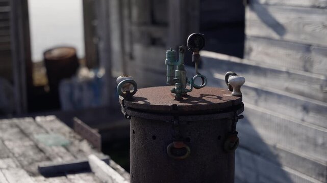 Weathered industrial cylinder with rusted valves and fittings on a sunlit wooden dock beside a coastal shack, showcasing patina, decay, and shadows in a waterfront setting.