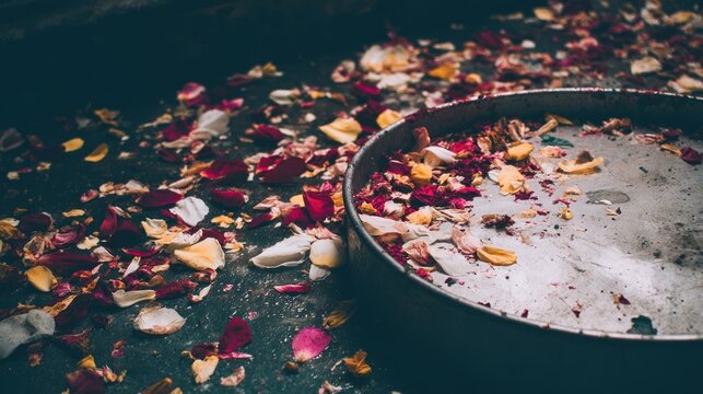 Scattered remnants of colorful flower petals cover a dark surface near a shallow metal container.