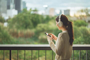 Young woman wearing headphones using smartphone on outdoor balcony, relaxing with music while...