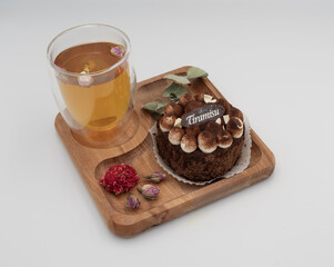 Tiramisu dessert served with herbal tea on a wooden tray, decorated with cocoa cream, dried roses and eucalyptus leaves. Cozy sweet treat on a clean white background.