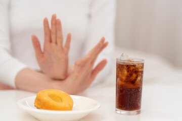 Woman on diet refusing soda and donut with hand gesture, symbolizing healthy lifestyle, sugar reduction, calorie control, and commitment to clean eating for weight loss.